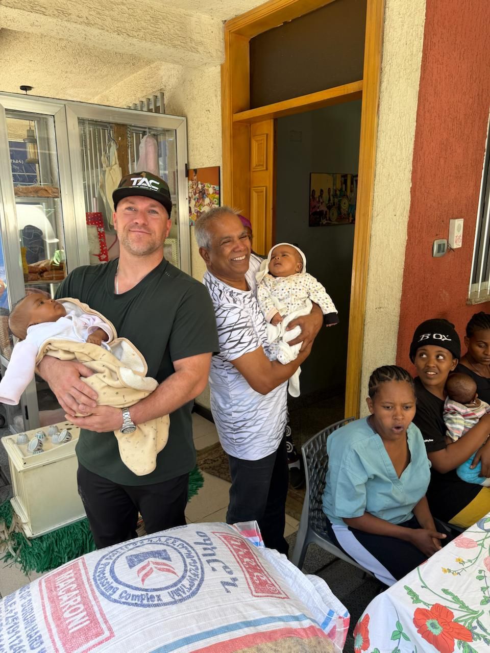 Two men each hold a baby while women sit nearby.